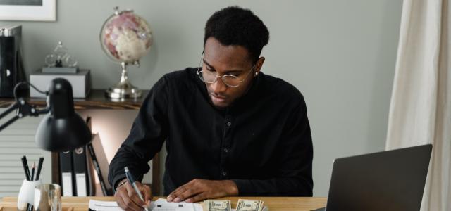 A young man sitting at a desk. There is a calculator and money on the desk, and the man is writing on a chart.