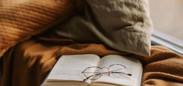 An open book on a windowsill. The book is resting on a knit blanket, and there is a pair of reading glasses on top of the book.